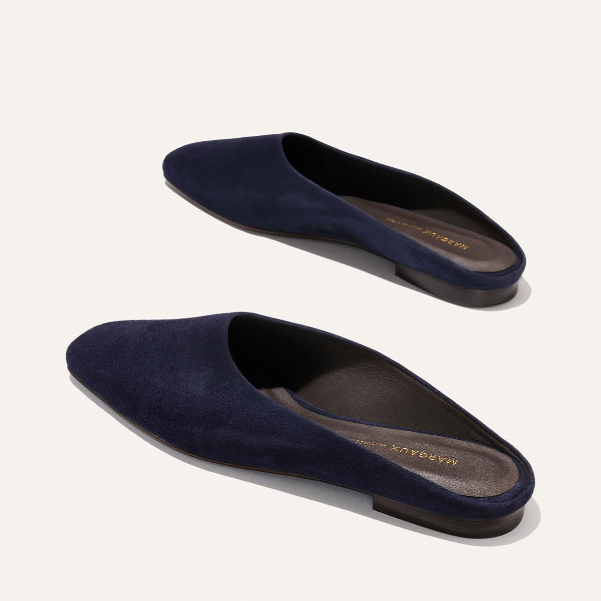 The Clara - Indigo Suede by Margaux: a pair of navy blue suede slip-on mules with flat brown soles and a slipper-like silhouette, shown on a plain off-white background.