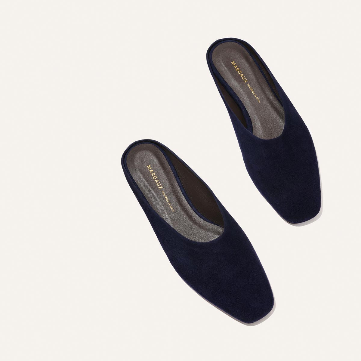 The Clara - Indigo Suede by Margaux features dark navy blue suede slip-on mules with a slipper-like silhouette, square toes, flat insoles with gold branding, and a versatile style perfect for any occasion.