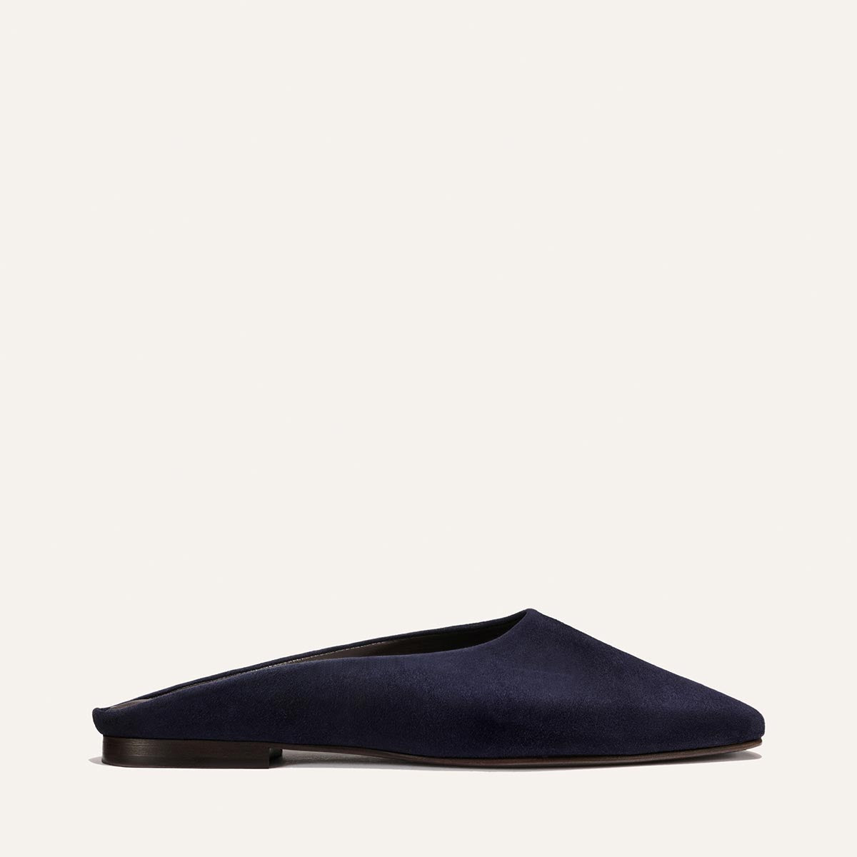 The Clara - Indigo Suede by Margaux is a dark blue suede mule with a closed, rounded toe, open back, flat silhouette, and low heel—delivering minimalist everyday style. Shown in profile against a light background.