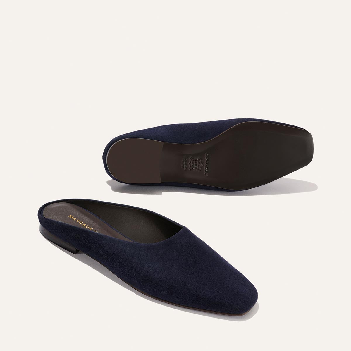 The Clara - Indigo Suede by Margaux: a pair of navy blue suede slip-on mules with a slipper-like silhouette and flat heel, one upright and one on its side revealing the sole and gold branding inside.