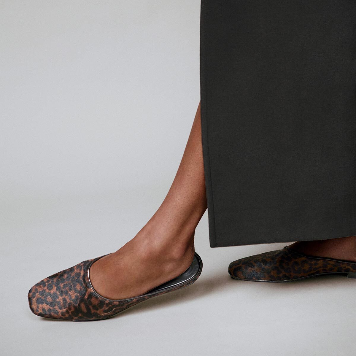 A person wearing a long dark skirt stands on a light background, with only the lower legs and feet visible, showcasing Margaux’s The Clara flats in Chocolate Leopard Haircalf.
