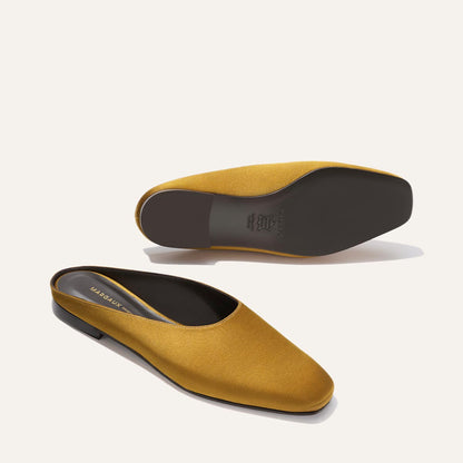 A pair of Margaux's The Clara mules in ochre satin, featuring black soles—one lying flat and the other turned to showcase the sole.