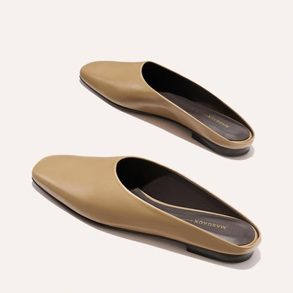 A pair of The Clara - Praline Calf leather slip-on mules by Margaux with flat black soles, shown top-down on a beige background. The dark-lined insole features "MARGAUX" printed in gold.