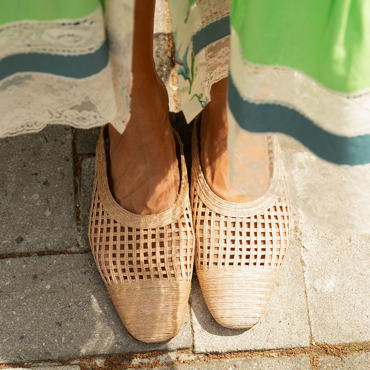 Someone stands on a stone pavement wearing Margaux's The Clara - Natural Grid Raffia shoes; part of a light green lace-trimmed skirt is also visible.