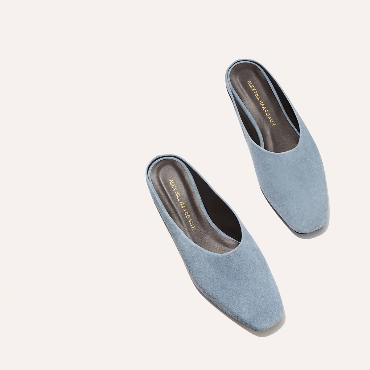 The Margaux Alex Mill x Margaux: The Clara - Slate Blue Suede mules, featuring pointed toes and low heels, are shown on a light beige background. Gold text details the insoles, accentuating their sleek, modern design and versatile style.