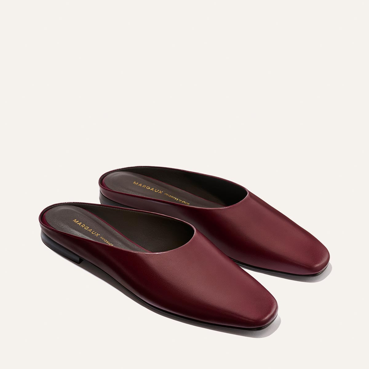 A pair of The Clara - Wine Calf slip-on mules by Margaux, crafted in rich burgundy leather with a subtle heel and displayed side by side on a neutral background, these shoes effortlessly blend slipper-like comfort with elevated chic for versatile, do-it-all style.
