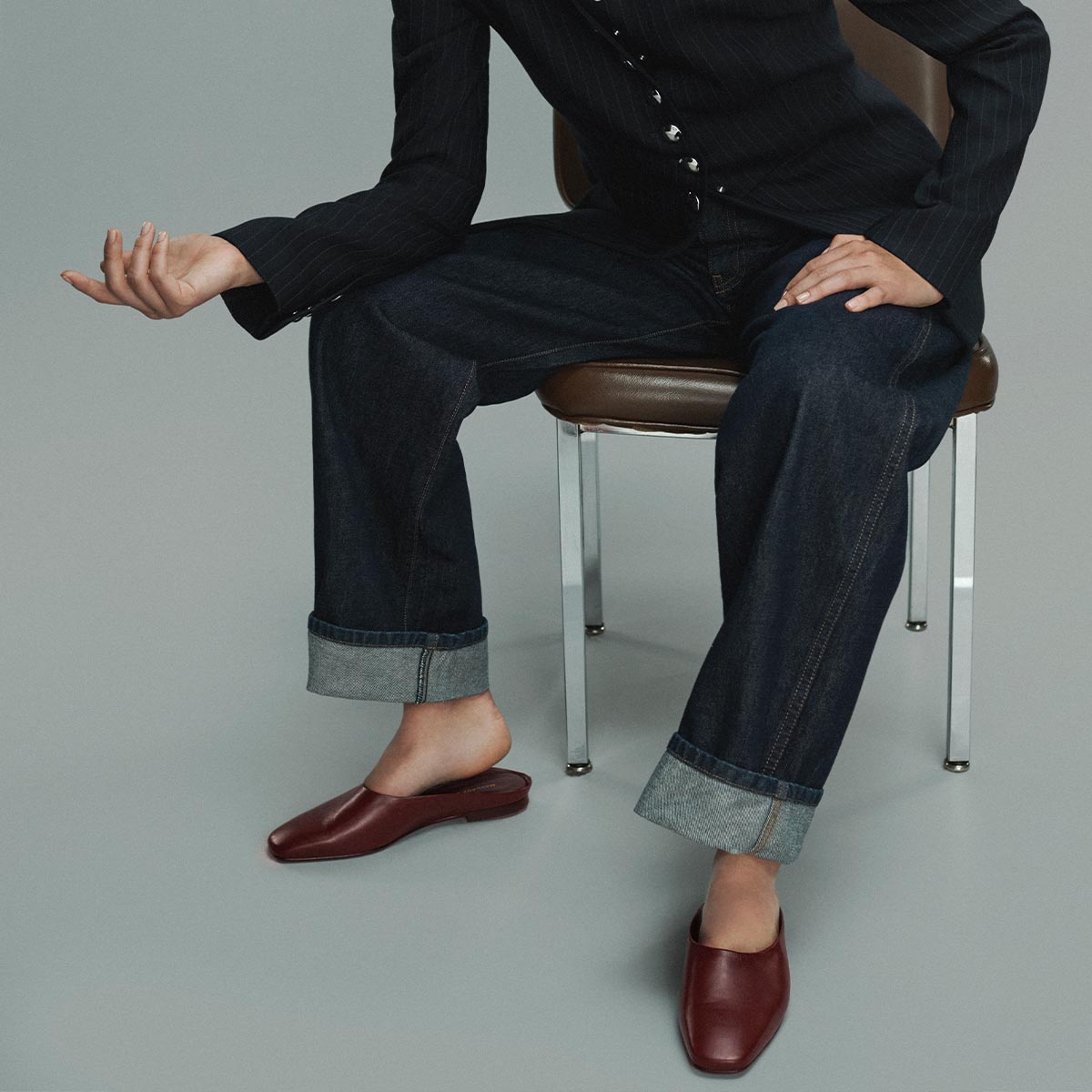 A person in dark jeans, a black jacket, and Margaux’s The Clara – Wine Calf slip-ons sits on a chair with one leg slightly raised and an arm bent, against a plain gray background.