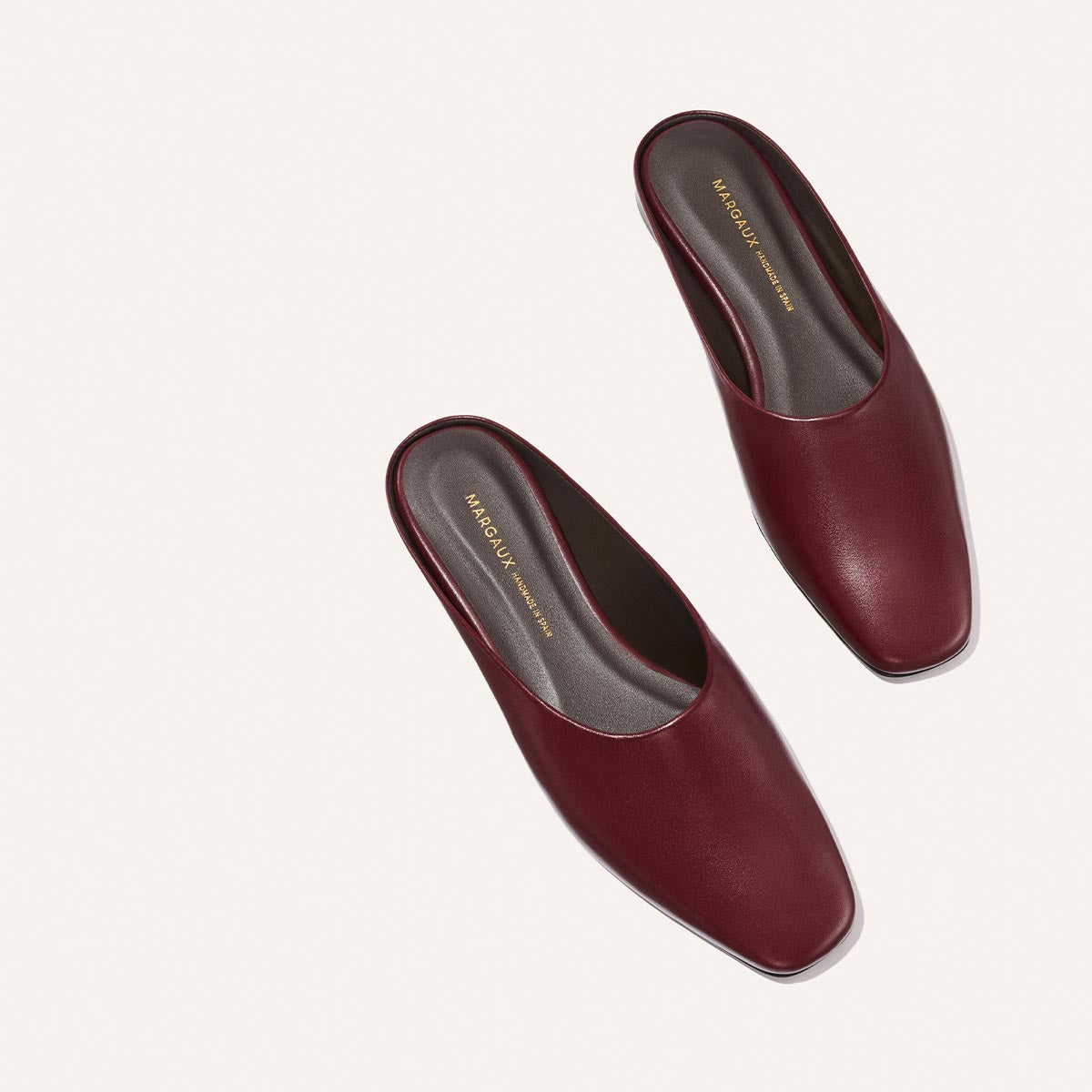 A pair of The Clara - Wine Calf slip-on mules by Margaux, featuring a square toe and versatile style, are displayed on a plain light background. The shoes have dark insoles with gold branding and appear new and unworn.