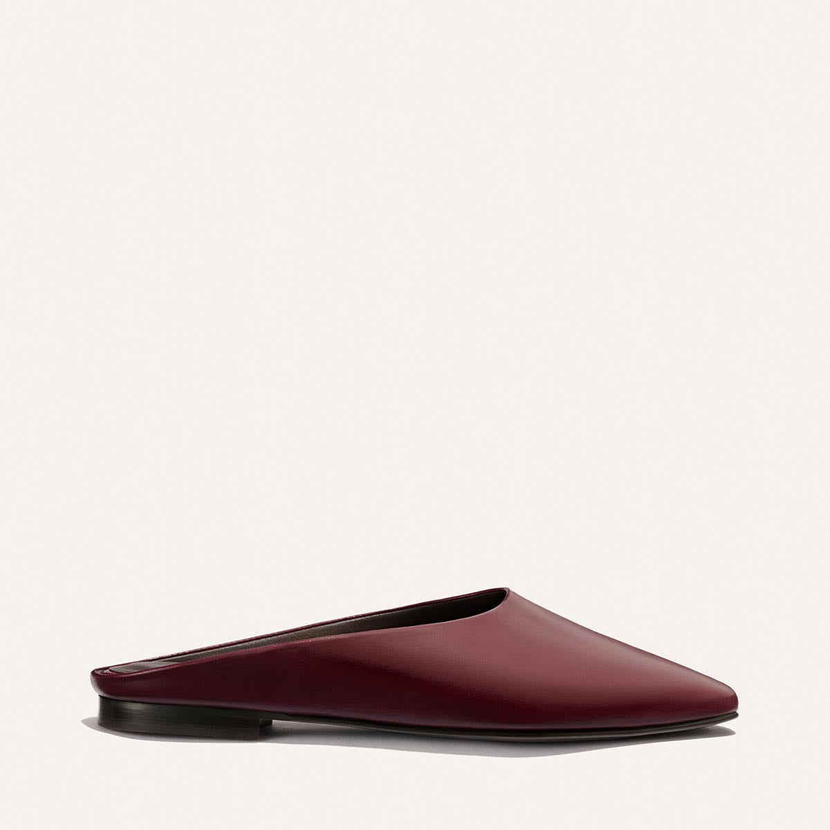 Introducing The Clara - Wine Calf by Margaux: a minimalist maroon leather mule with a closed, pointed toe and flat black sole, showcasing a slipper-inspired silhouette and set against a simple, light background.