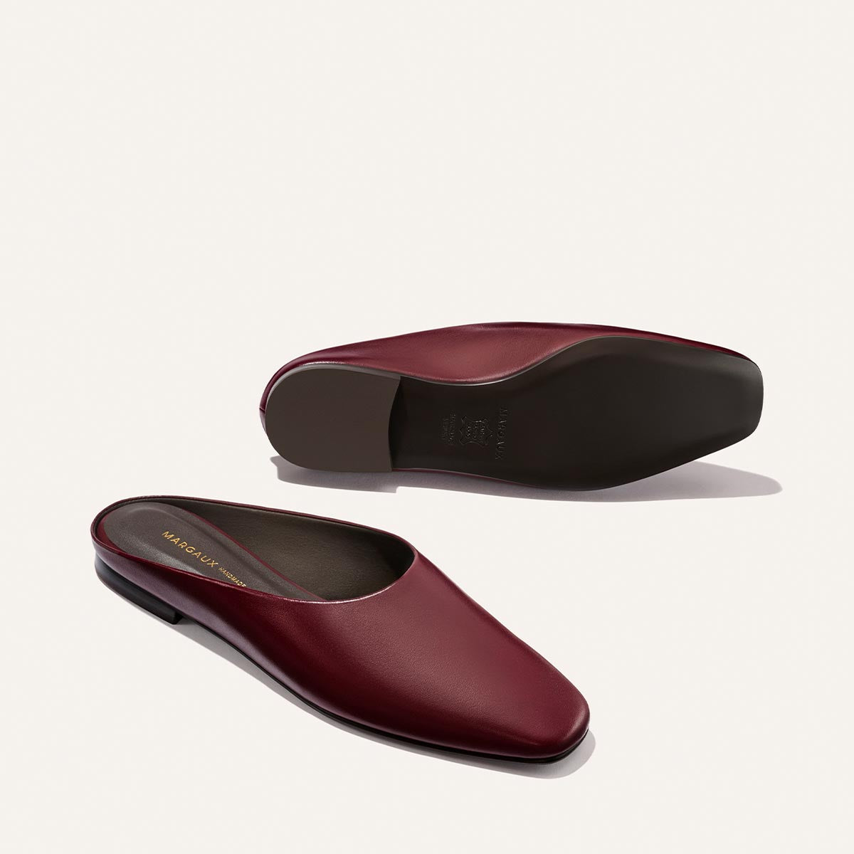 A pair of Margaux's The Clara - Wine Calf leather slip-on flats with a slipper silhouette, closed toes, and open backs, displayed on a light background. One shoe stands upright while the other lies on its side to showcase the sole.