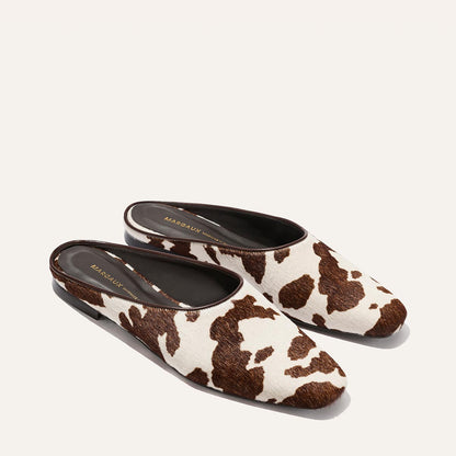 The Clara - Cow Print Haircalf by Margaux are slip-on mules with a brown and white cow print, flat sole, black lining, and "Margaux" printed in gold inside.