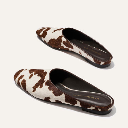 The Clara - Cow Print Haircalf by Margaux are slip-on mule shoes with a brown and white cow print, black soles, dark brown lining, and “Margaux” gold-printed on the insole.