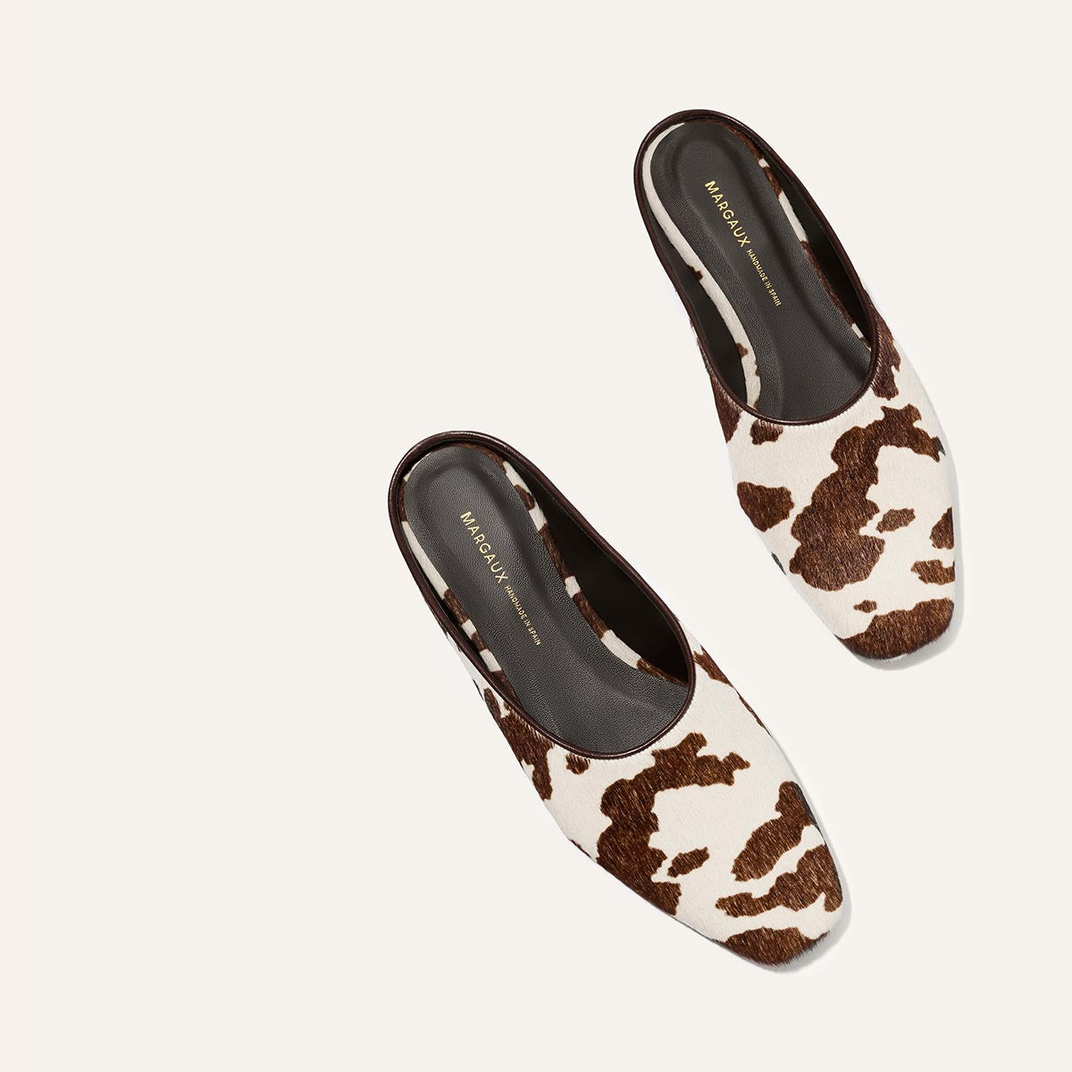 The Clara - Cow Print Haircalf by Margaux: women's slip-on mule shoes with a brown and white cow print pattern, black insoles, and low heels, displayed on a plain light background.