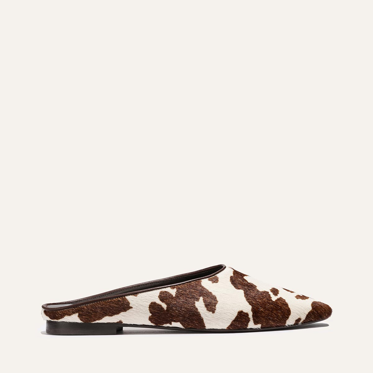 The Clara - Cow Print Haircalf by Margaux is a slip-on mule with a flat heel, featuring a brown and white cow print pattern, shown against a light background.