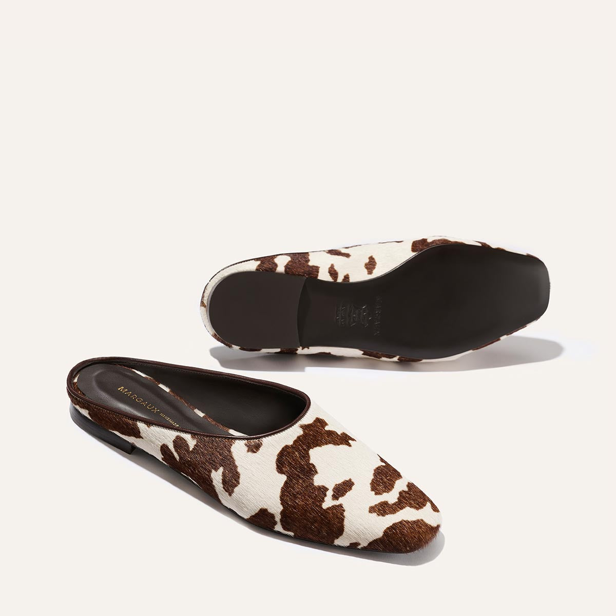 Margaux’s The Clara - Cow Print Haircalf slip-on flats feature a brown and white cow-print pattern, with one shoe displayed flat and the other on its side to reveal the black sole.