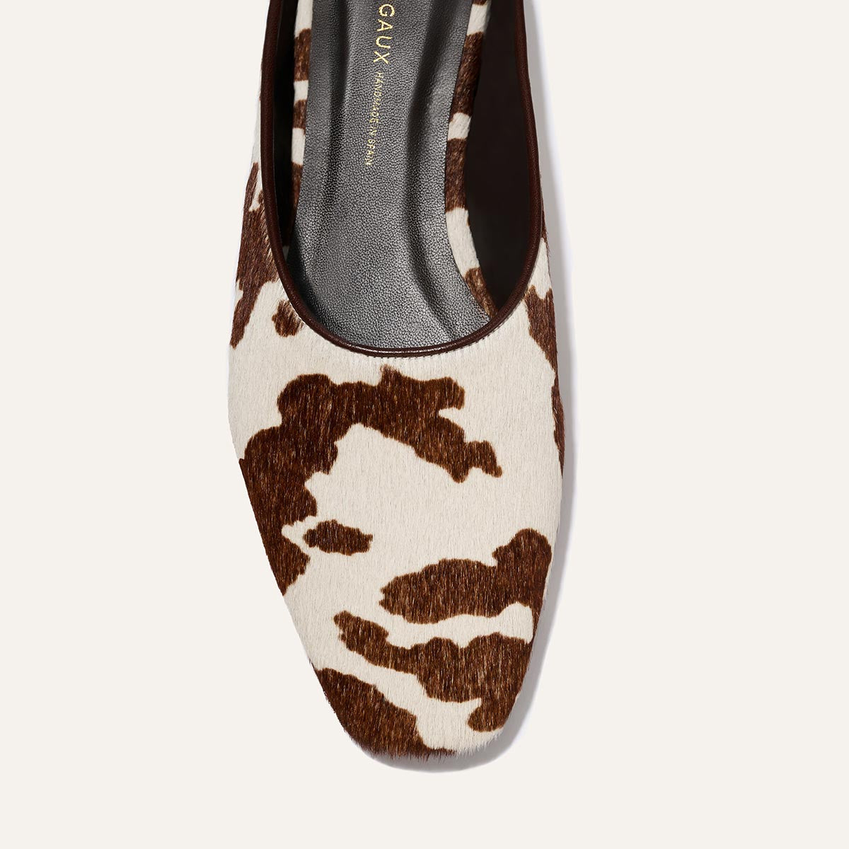 A close-up top view of Margaux's The Clara - Cow Print Haircalf flat shoe featuring a brown and white cow-print pattern, black insole, and rounded square toe.