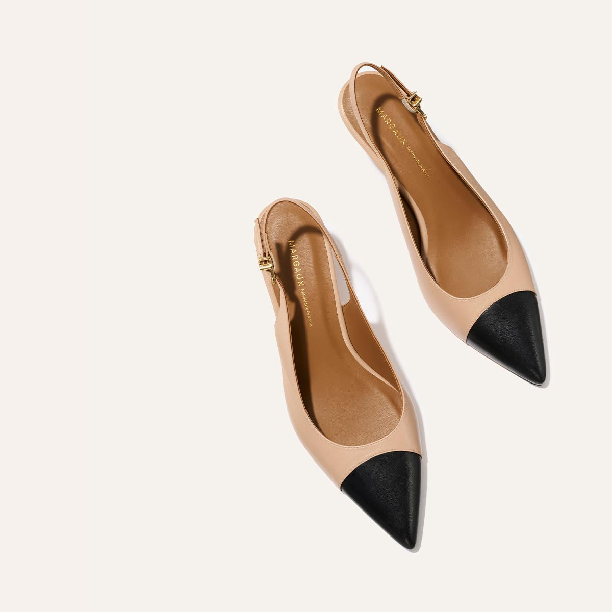 The Margaux Colette in Dune and Black Nappa: beige slingback flats with black snip toe, gold buckle accents, and a subtle micro heel, displayed on a light background.
