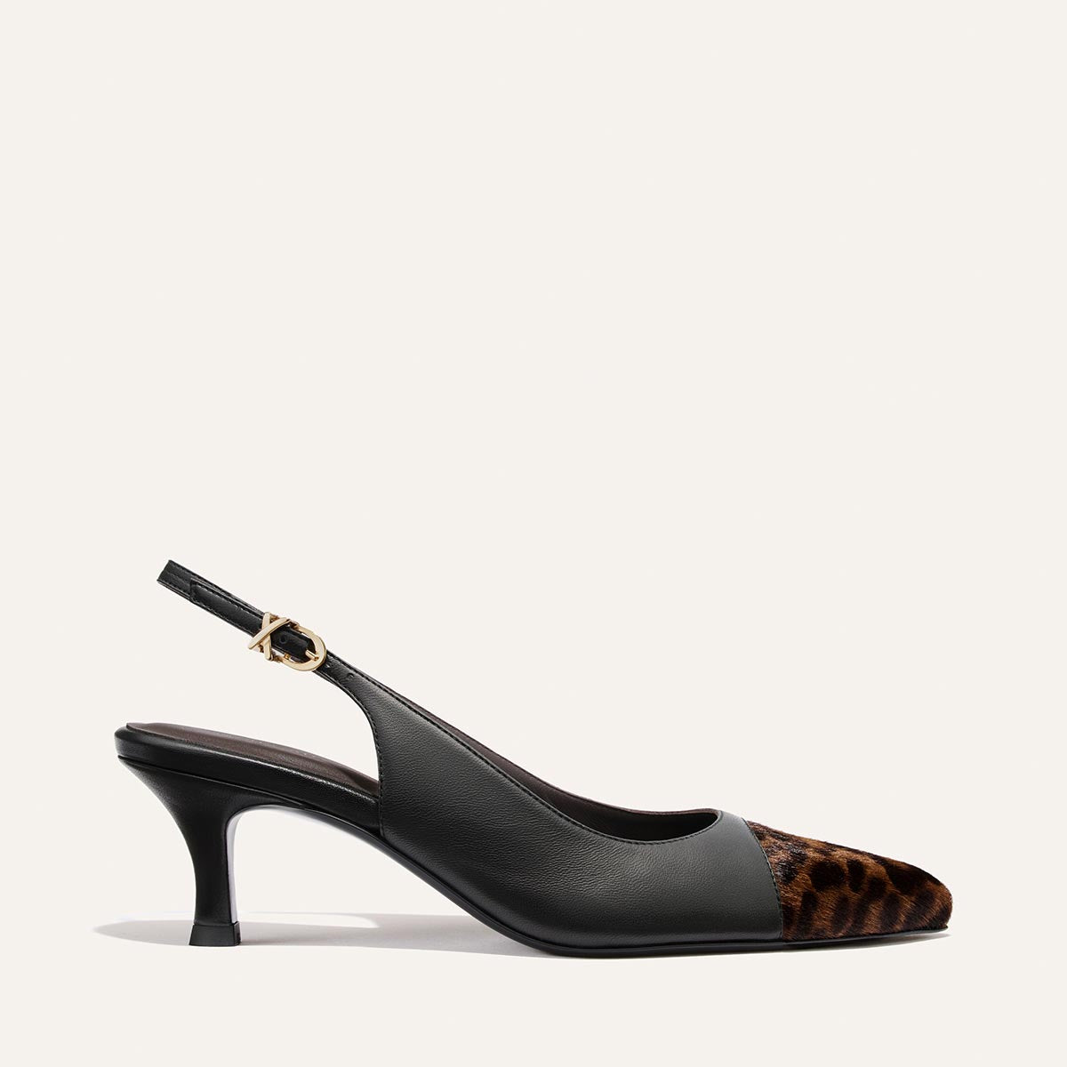 Margaux’s The Colette in Black Nappa and Leopard Haircalf is a black slingback heel with a micro heel and snip nose, featuring a leopard print front, leather upper, and a gold buckle on the ankle strap, set against an off-white background.