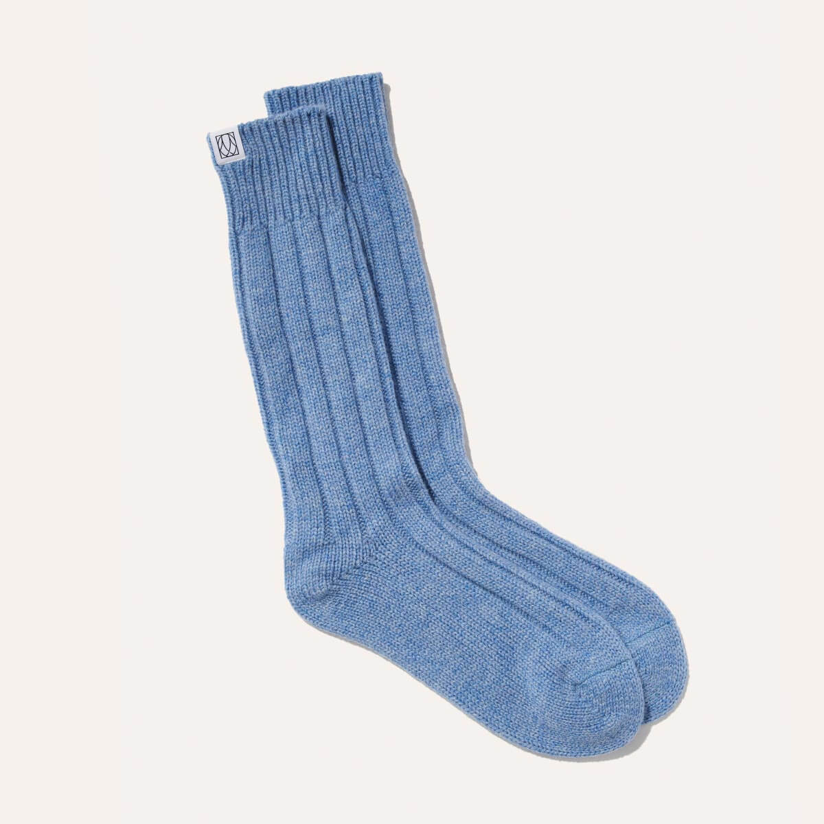 The Cozy Sock by Margaux is a light blue, ribbed crew sock made from a soft wool-cashmere blend, featuring a white tag with a blue logo on top—perfect for luxurious lounging. Shown on a plain white background.