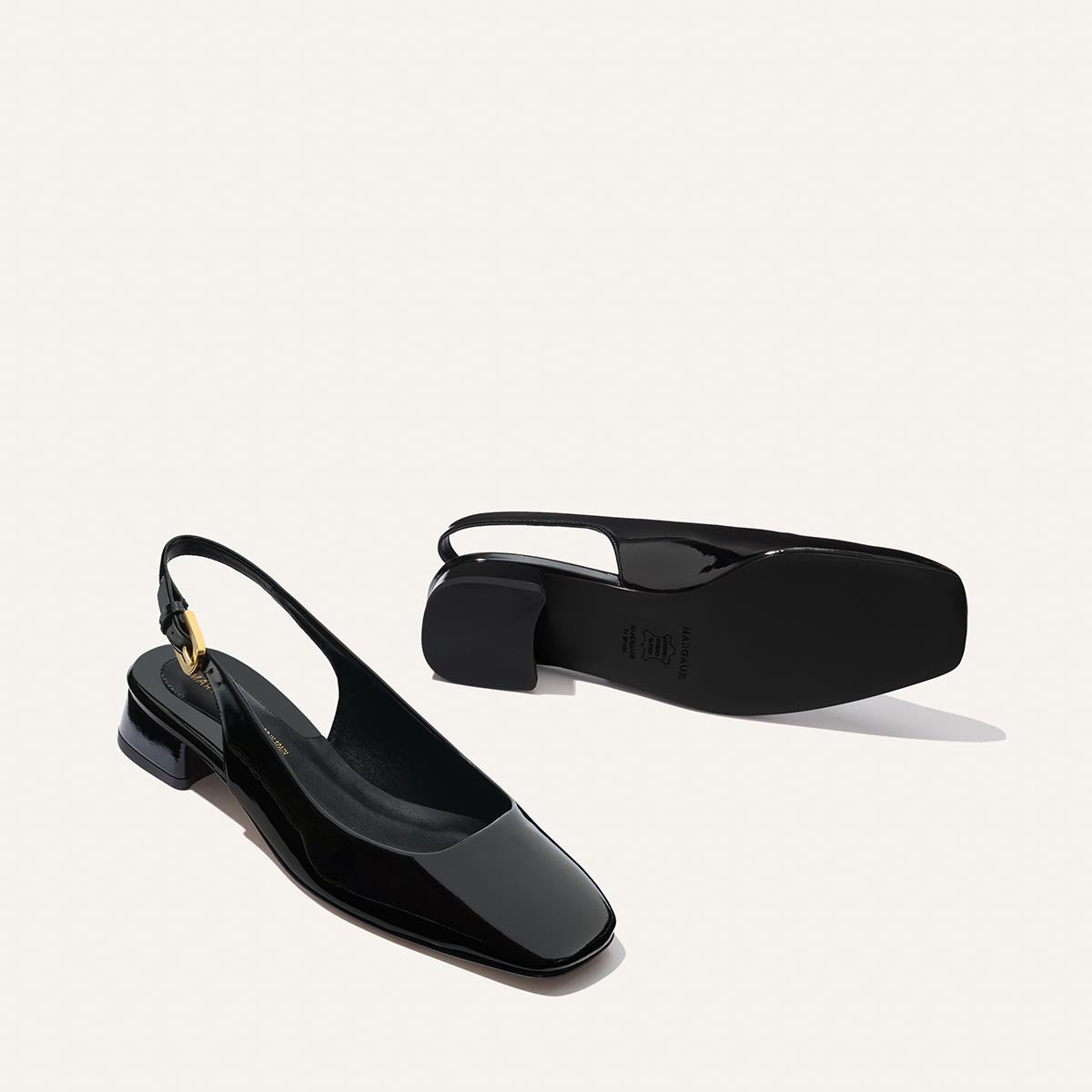 The Darcy Slingback - Black Patent by Margaux: sleek black patent leather ballet pumps with low block heels and square toes. Displayed on a light background, one shoe stands upright while the other lies on its side to show the sole.