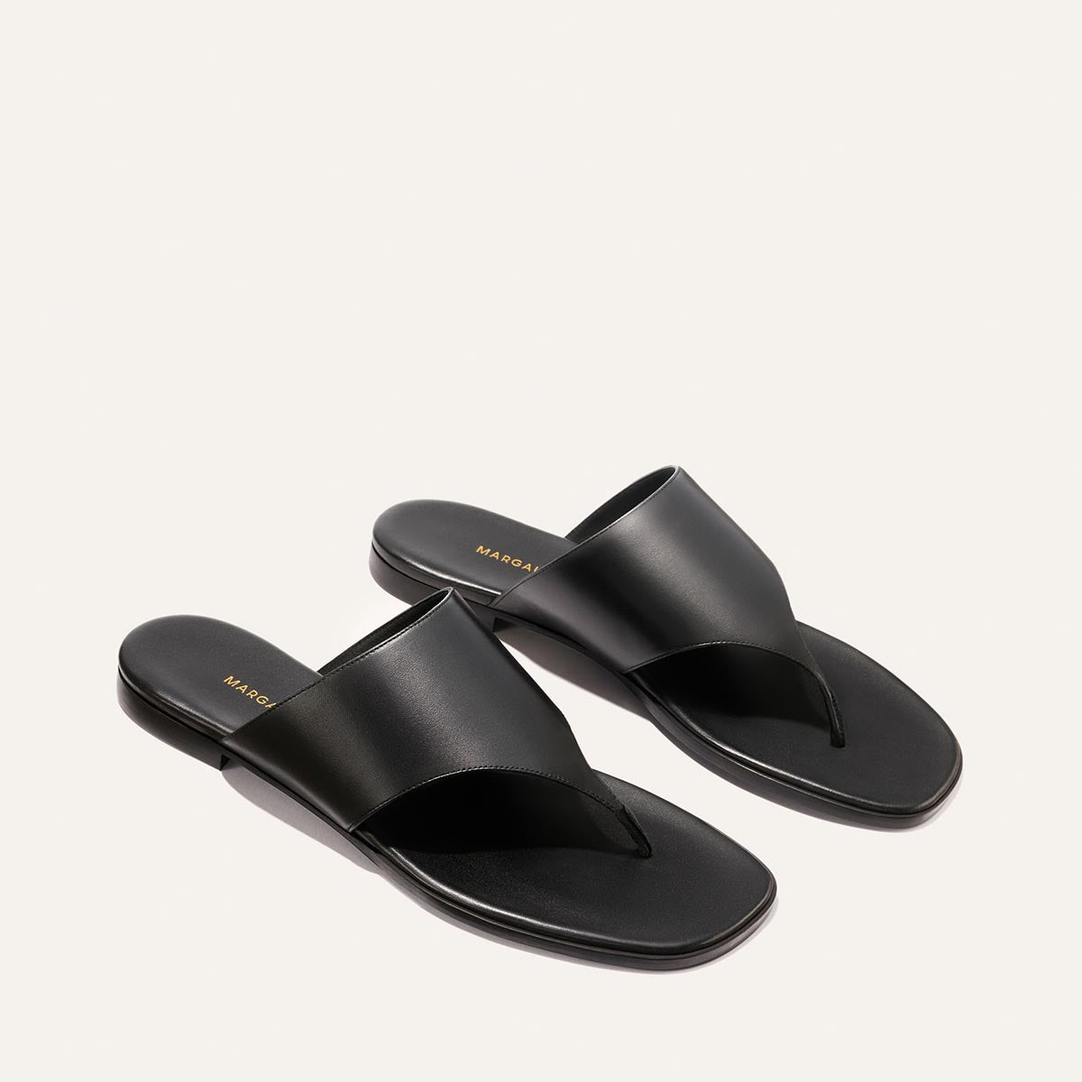 The Deia - Black Nappa by Margaux: black leather thong sandals with a wide upper strap and flat soles, shown on a light beige background.