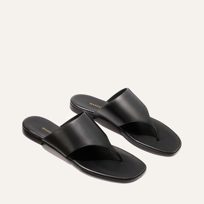 The Deia - Black Nappa by Margaux: black leather thong sandals with a wide upper strap and flat soles, shown on a light beige background.