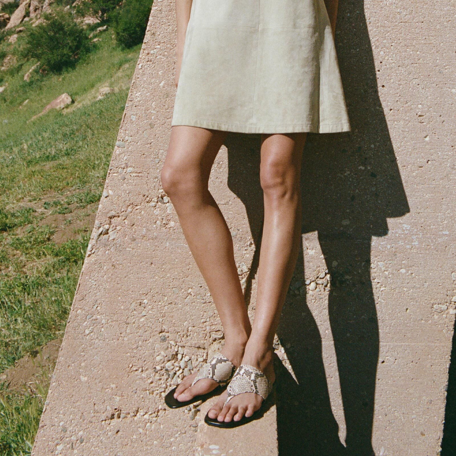 A person wearing a light-colored dress and Margaux's The Deia - Natural Python Embossed sandals stands outdoors by a slanted concrete wall, with grass and rocks in the background.
