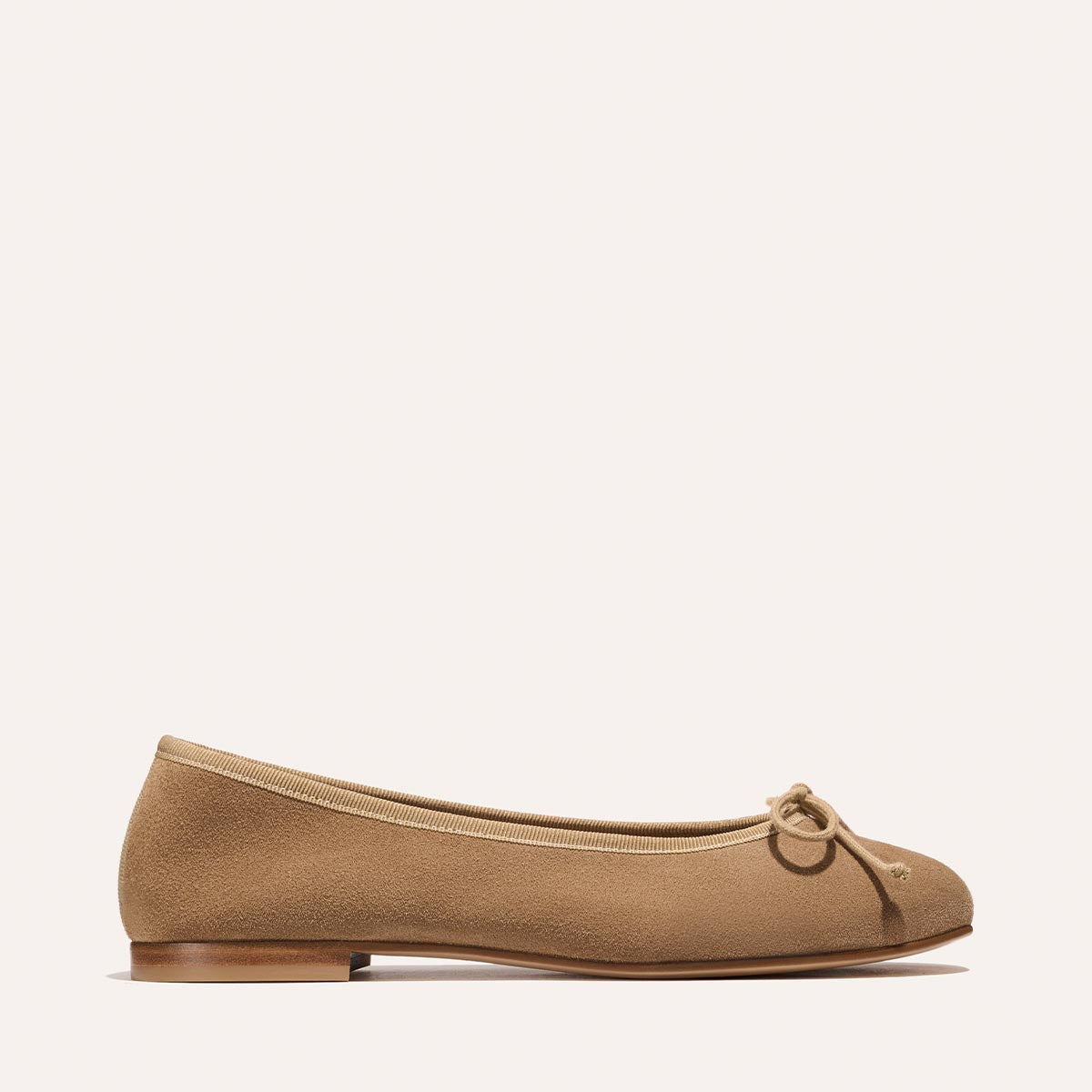 The Demi - Biscotti Suede by Margaux is a tan suede ballet flat, handmade in Spain, featuring a rounded toe, decorative bow, and small flat heel, shown in profile against a plain light background.