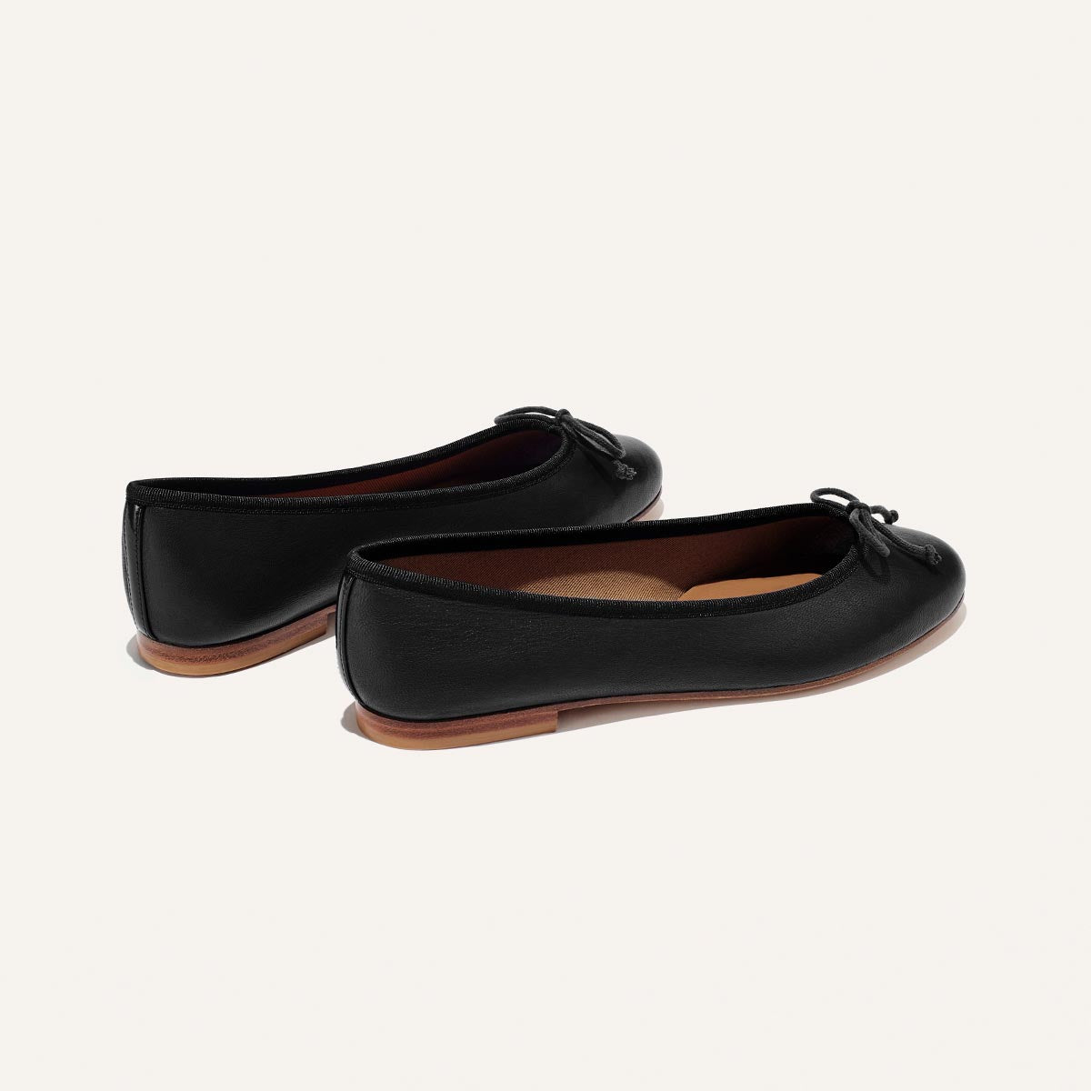 The Demi - Black Nappa from margaux-development are handmade black ballet flats with small bows, brown soles, and a classic design, crafted in Spain and shown on a plain light background.