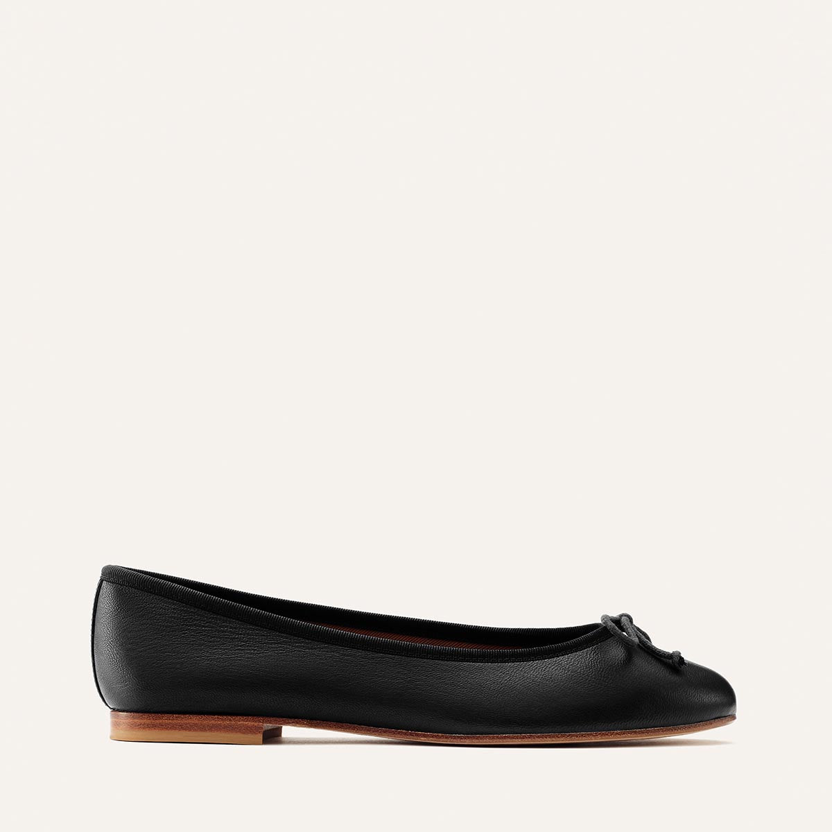 The Demi - Black Nappa by margaux-development is a handmade black leather ballet flat from Spain, featuring a small bow on the toe and a brown flat sole, shown against a plain white background.