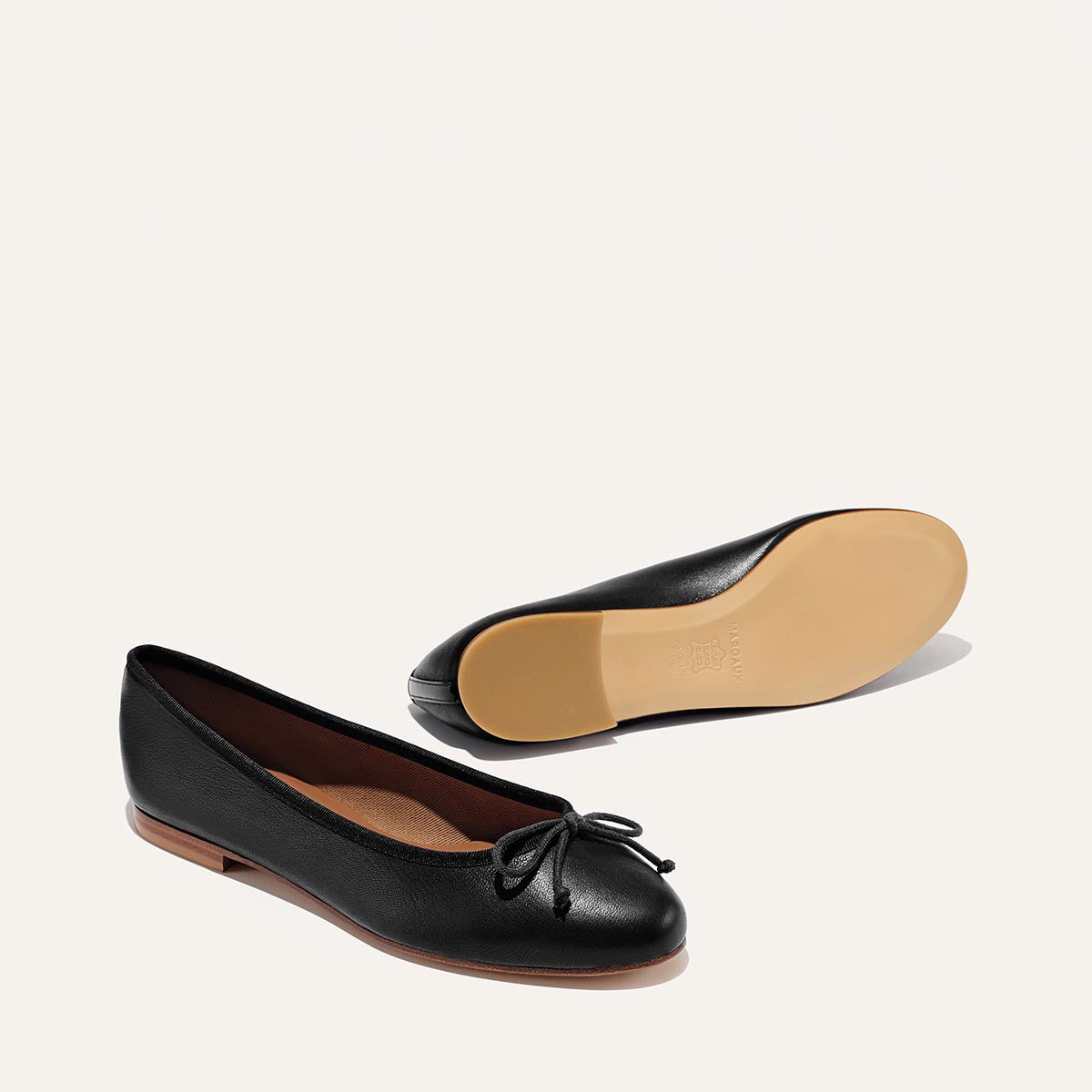The Demi - Black Nappa by margaux-development: black leather ballet flats handmade in Spain, featuring a small bow. One shoe shows the upper and bow; the other reveals the beige sole. Both are displayed on a plain white background.