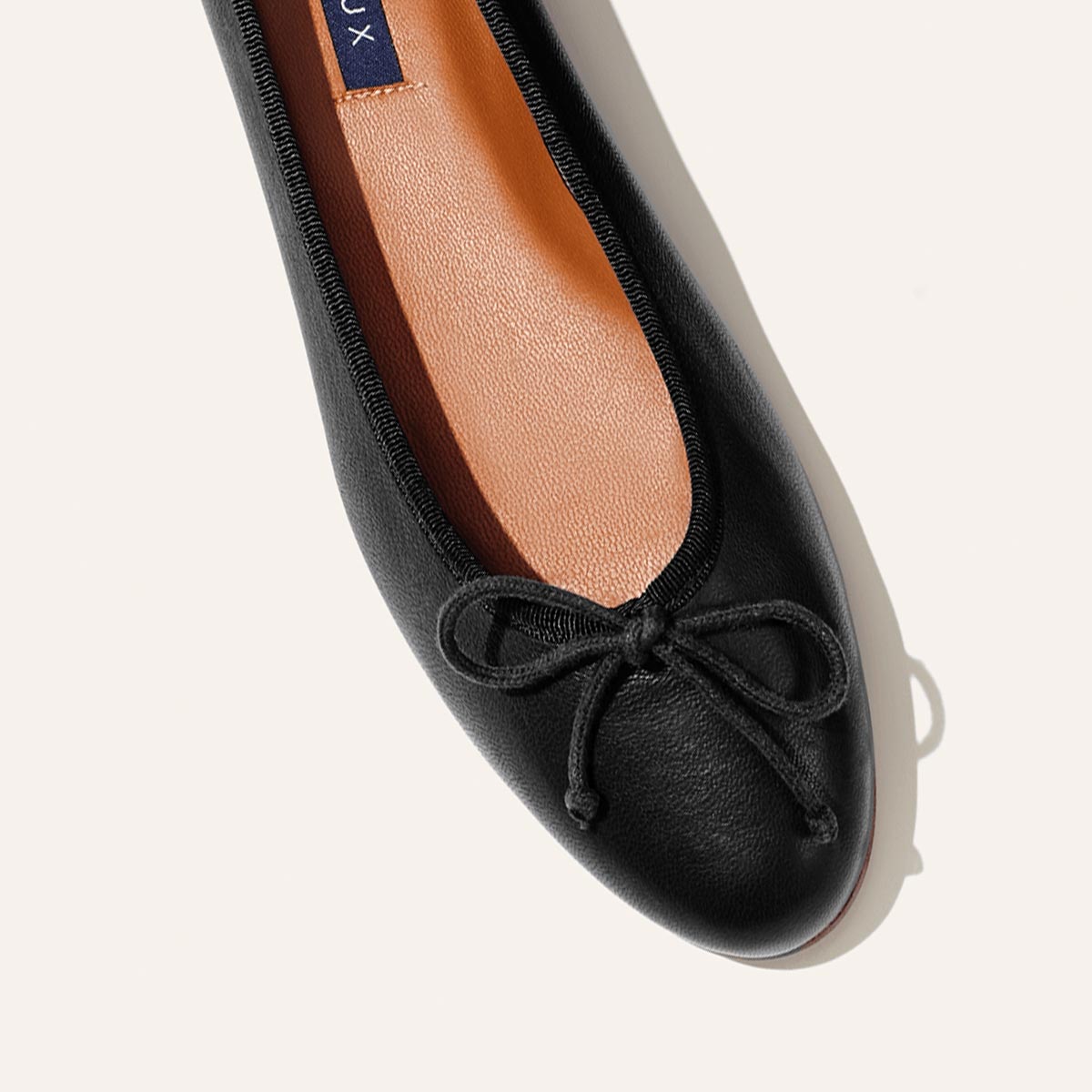 A close-up of The Demi - Black Nappa by margaux-development, a handmade Spanish ballet flat with a small bow on the toe and a tan insole, shown against a light background.