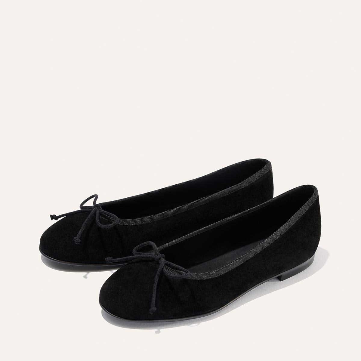 A pair of The Demi - Black Suede ballet flats by margaux-development, handmade in Spain with round toes and small bows on the front, displayed on a plain white background.