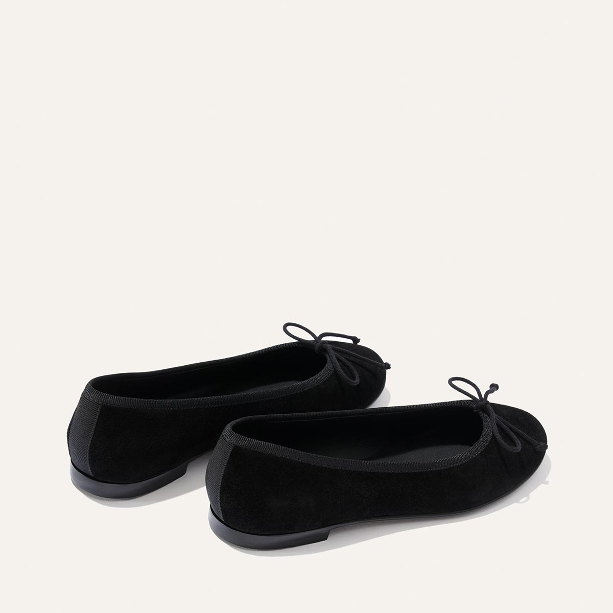 A pair of The Demi - Black Suede ballet flats by margaux-development, handmade in Spain from soft black suede and adorned with delicate bows at the toes, are displayed on a light background, with one comfortable shoe positioned just in front of the other.