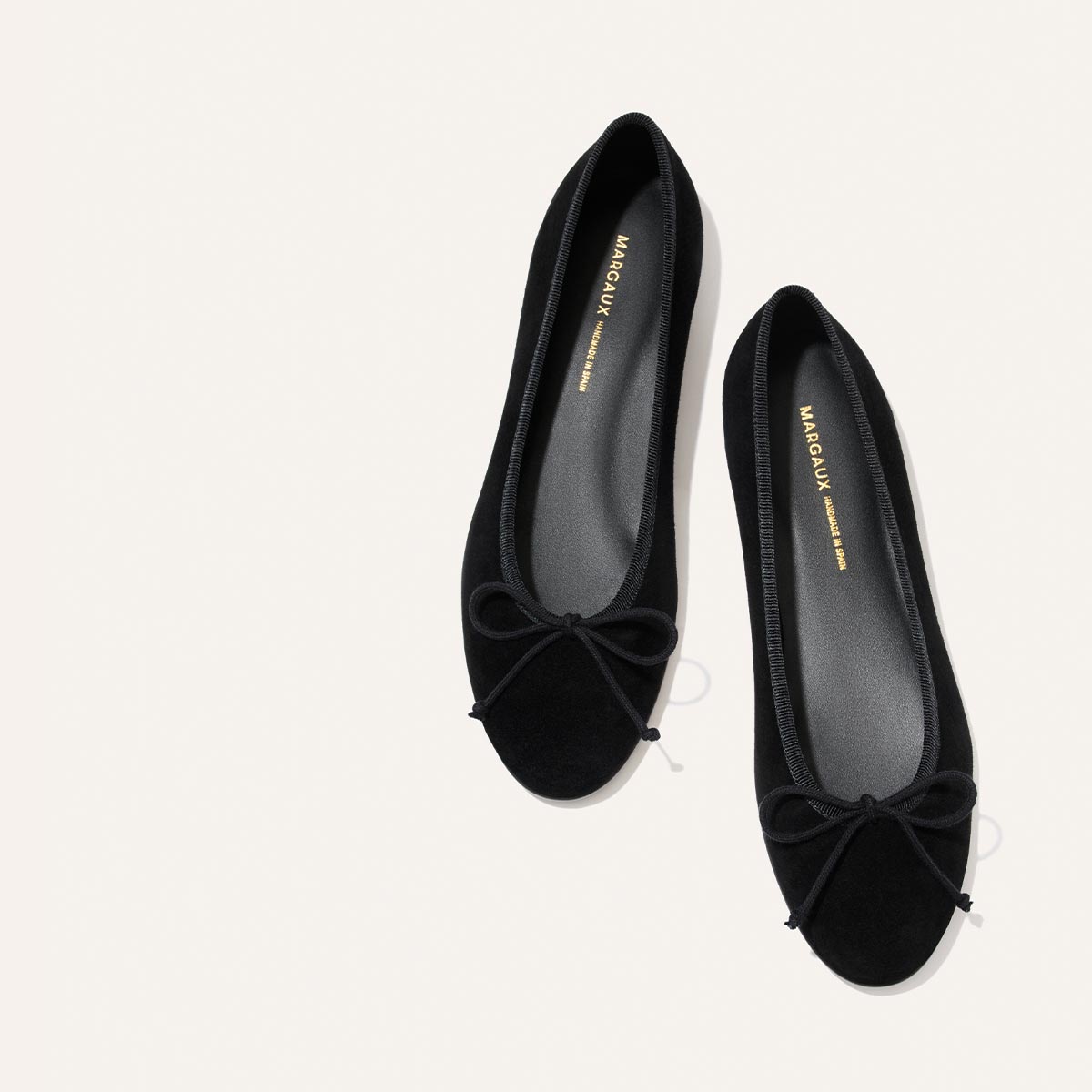 A pair of The Demi - Black Suede ballet flats by margaux-development, featuring small bows on the toes, is displayed against a plain light background. Handmade in Spain, these comfortable shoes include insoles labeled MARGAUX.