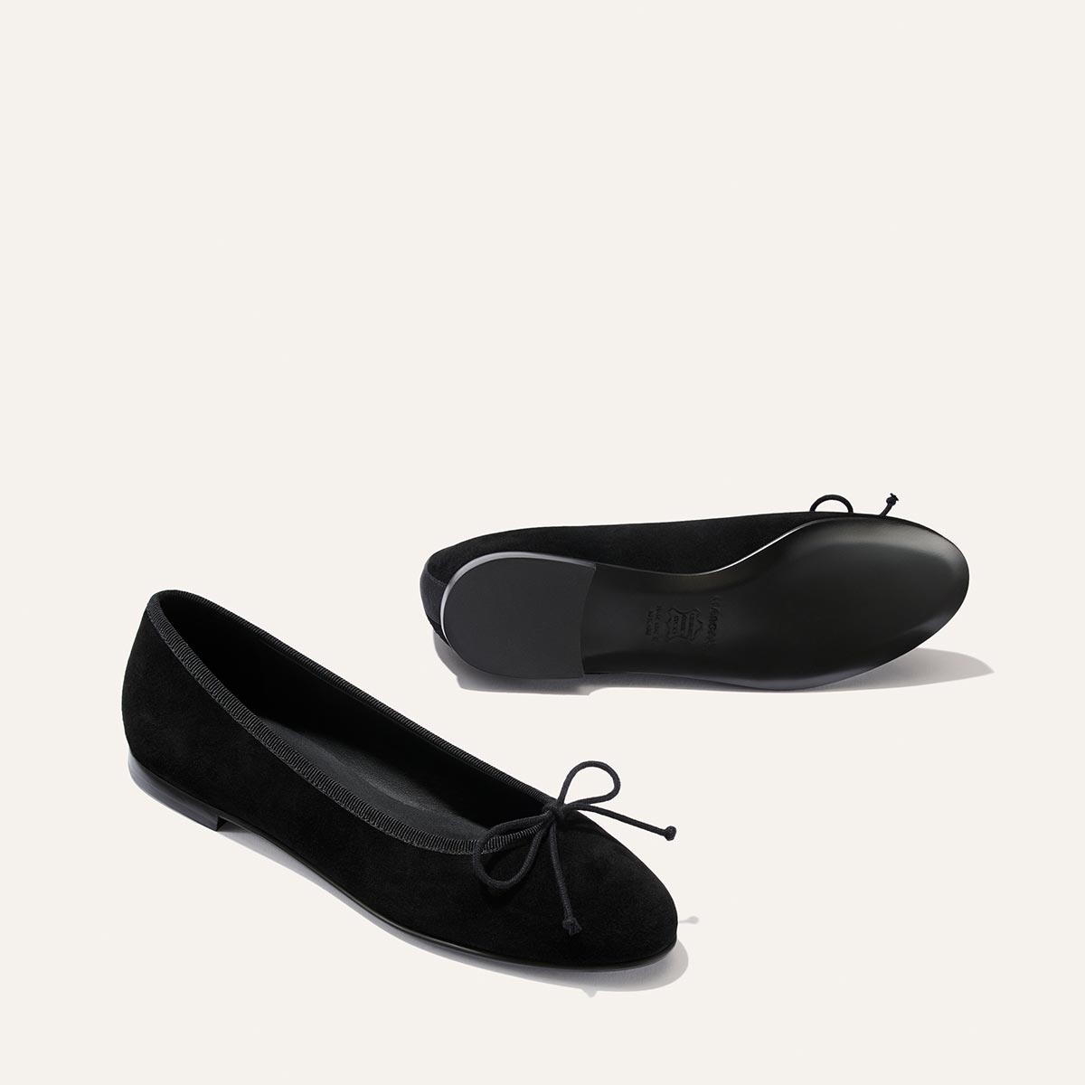 A pair of The Demi - Black Suede ballet flats by margaux-development, handcrafted in Spain from black suede and adorned with petite bows on the toes, is displayed against a plain light background. One comfortable shoe stands upright, while the other lies on its side to reveal the sole.