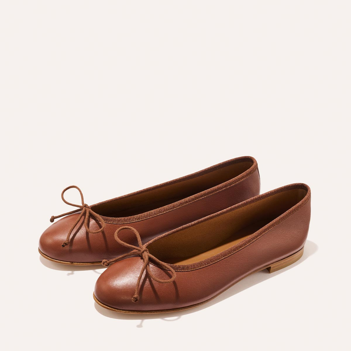The Demi - Cognac Nappa by margaux-development is a handmade Spanish ballet flat in brown, featuring rounded toes, a small bow, and flat soles, shown on a light background.