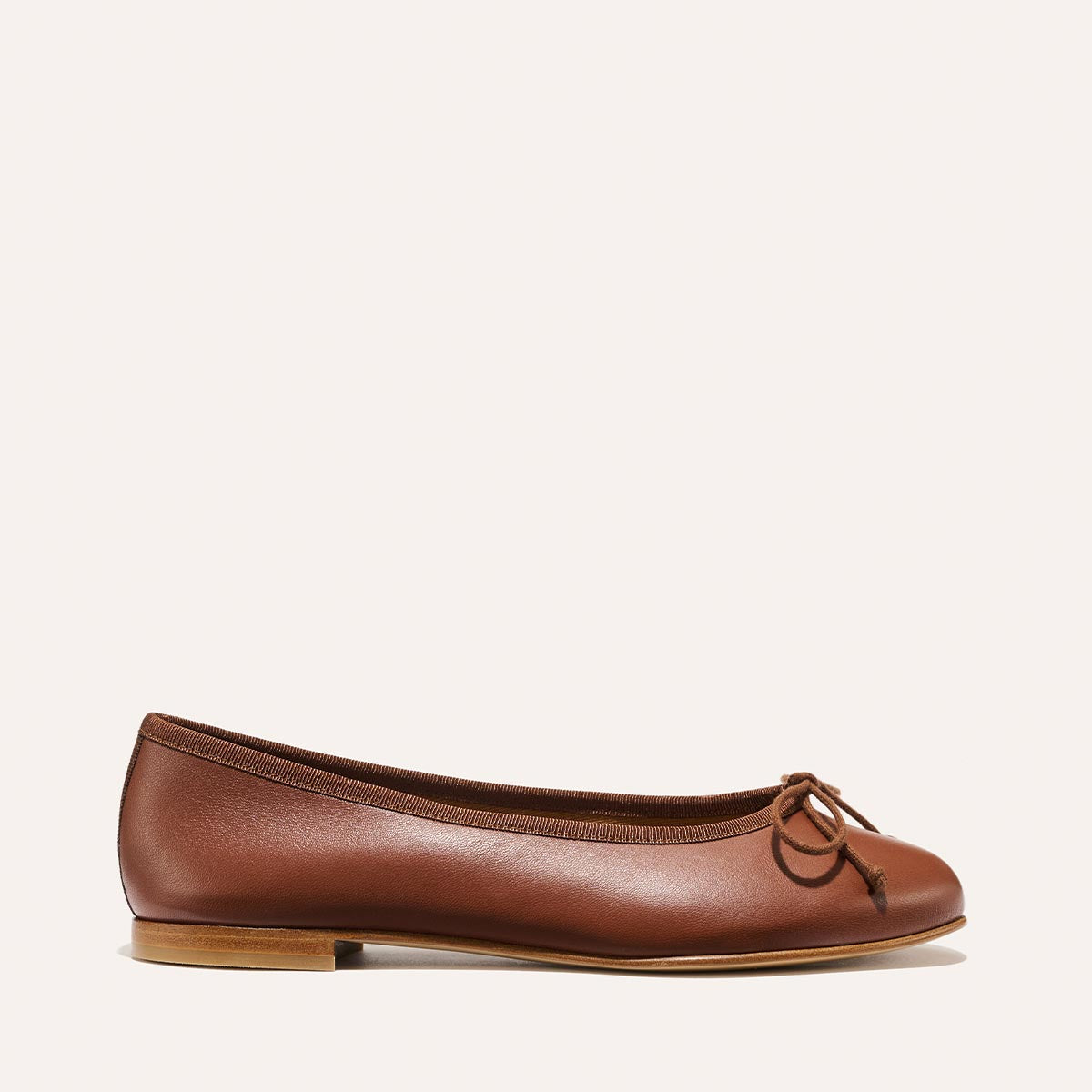 The Demi - Cognac Nappa by margaux-development is a brown leather ballet flat handmade in Spain, featuring a small bow, low wooden heel, and rounded toe, displayed on a plain light background.