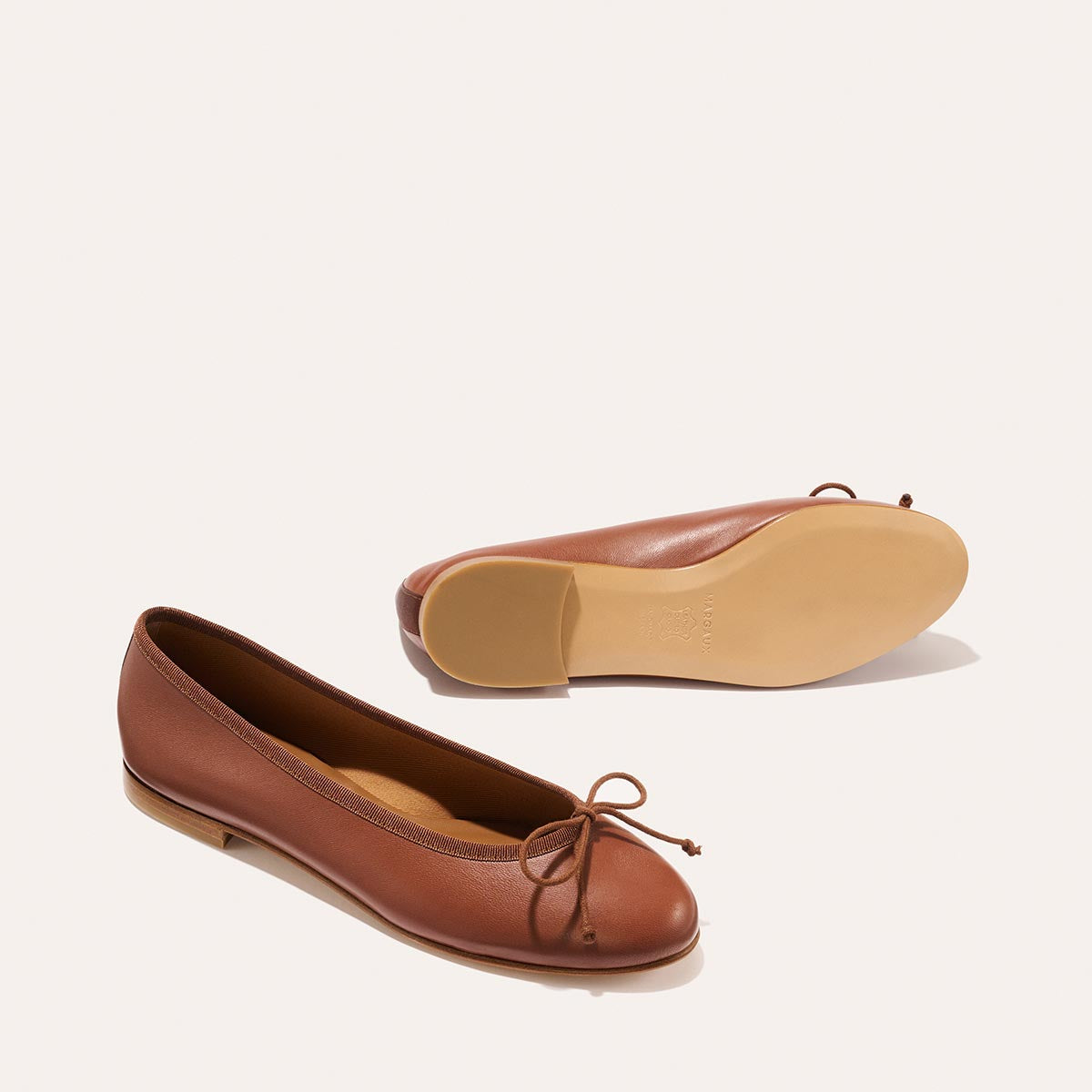 The Demi - Cognac Nappa by margaux-development: a pair of brown leather ballet flats, handmade in Spain with small toe bows—one upright, the other on its side to reveal the sole—displayed on a light background.