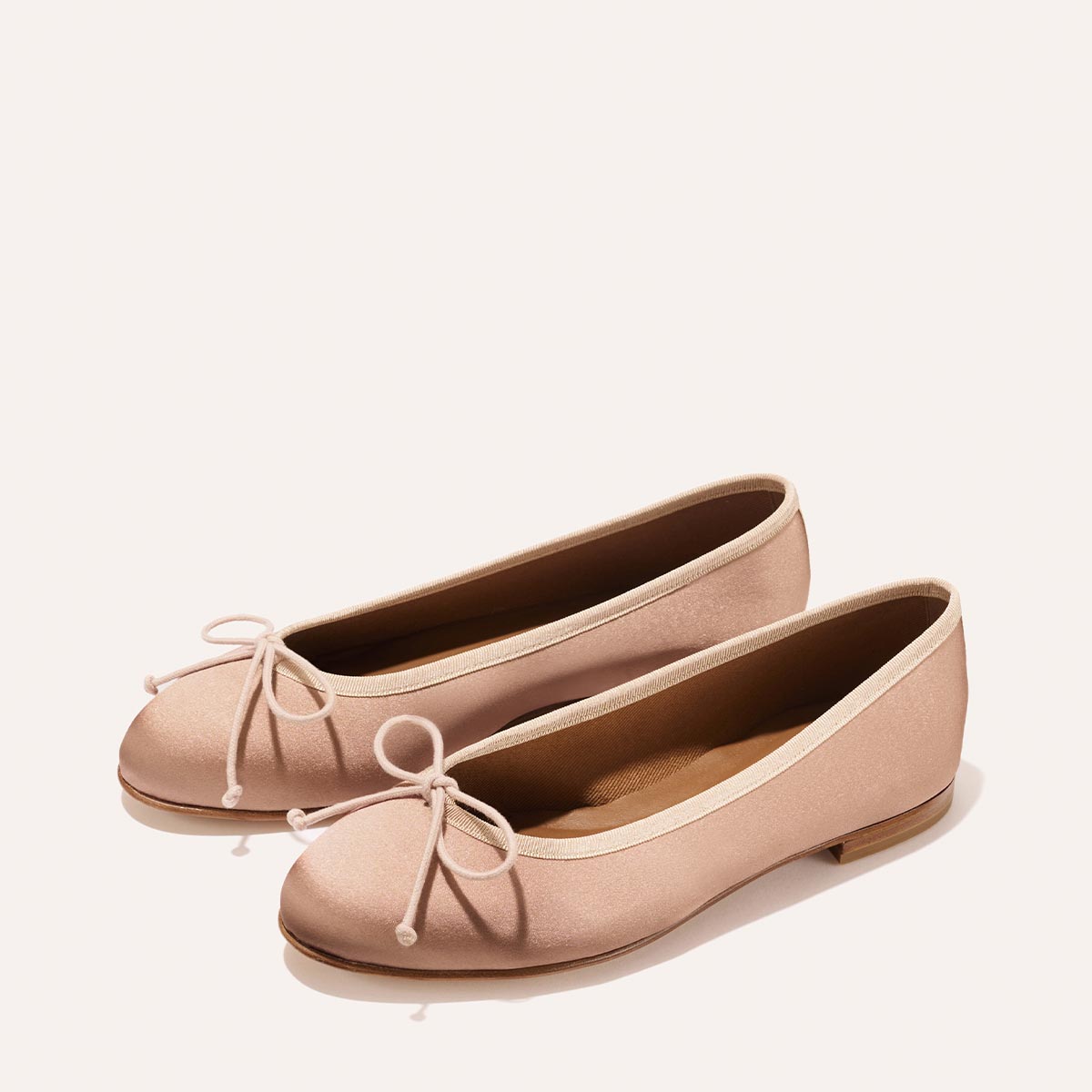 The Demi - Dusty Pink Satin ballet flats by margaux-development, handmade in Spain, feature rounded toes, matching front bows, and light brown soles—shown on a plain off-white background.
