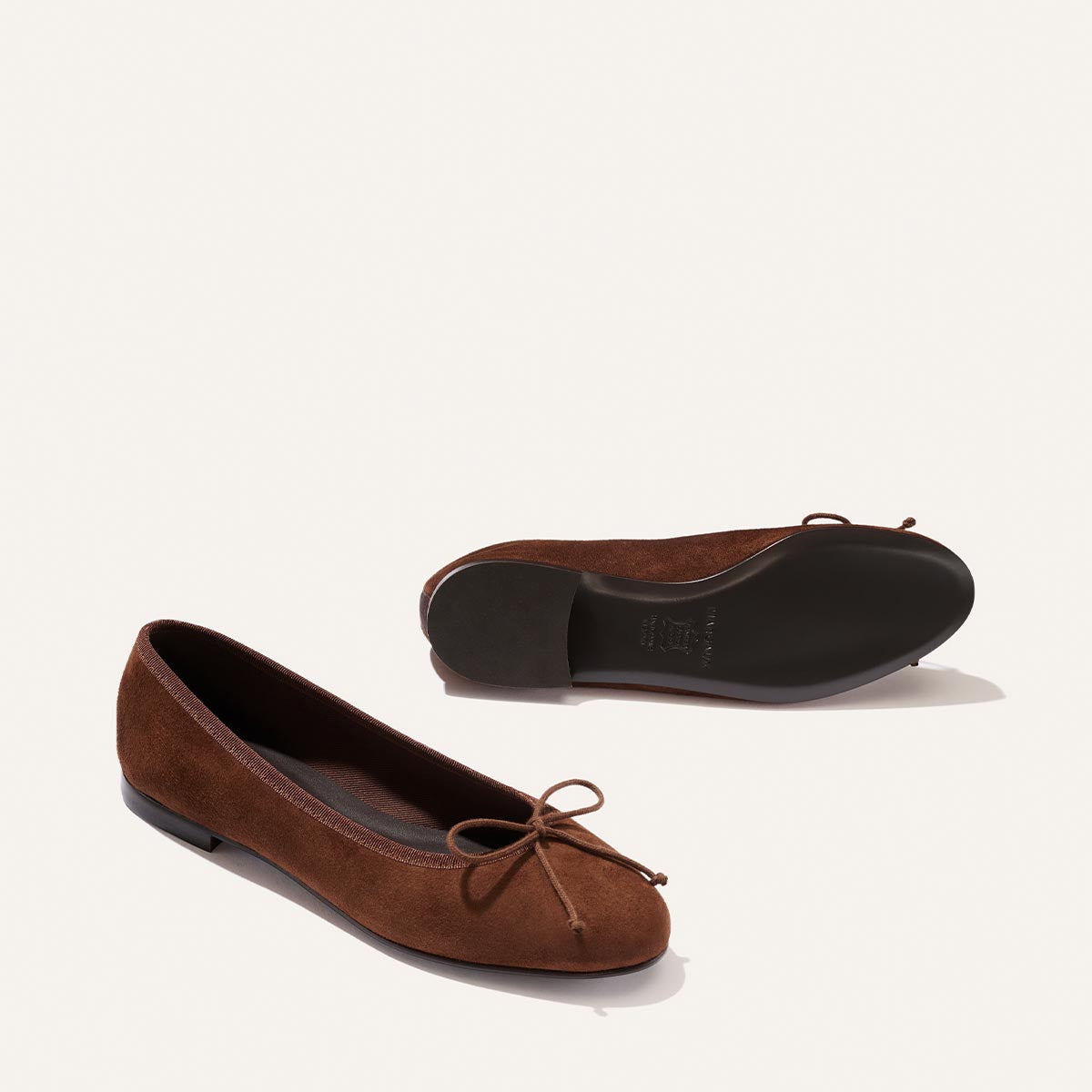 A pair of Margaux's The Demi - Ganache Suede ballet flats, handmade in Spain from rich brown suede, are displayed on an off-white background. One shoe stands upright while the other lies on its side to reveal the black sole; both feature delicate bows at the toes.