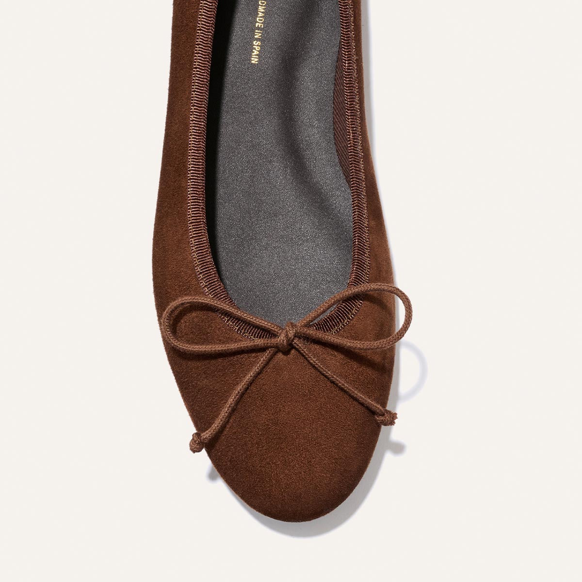 A close-up of The Demi – Ganache Suede ballet flat by Margaux, a comfortable brown suede shoe with a bow on the toe, handmade in Spain and shown from above on a plain light background.