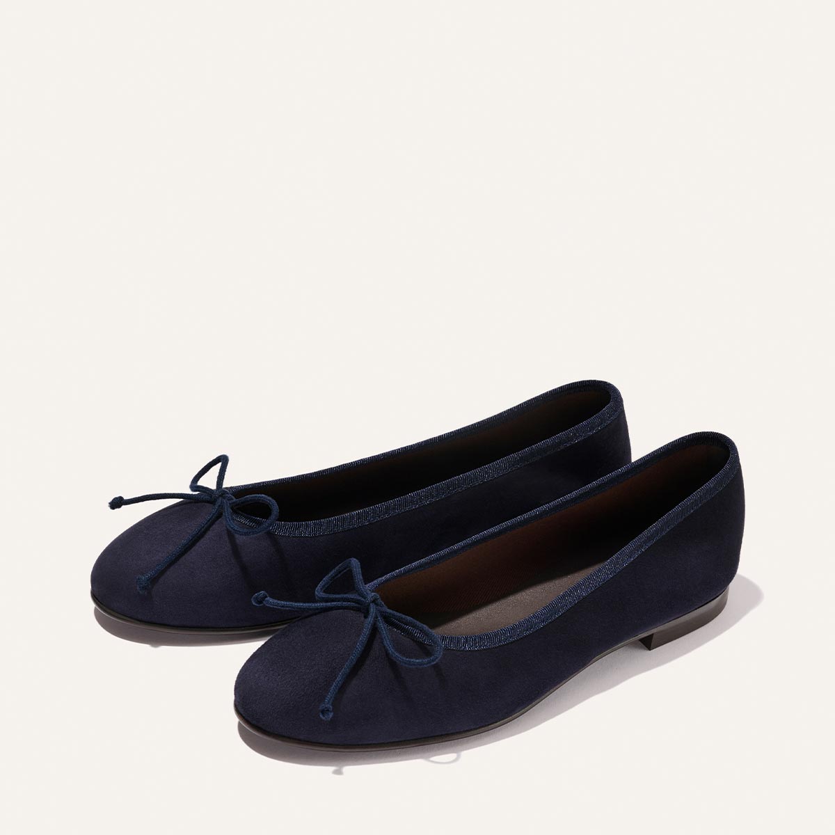 The Demi - Indigo Suede by Margaux are comfortable navy blue ballet flats with round toes and decorative bows, handcrafted in Spain and shown on a plain, light background.