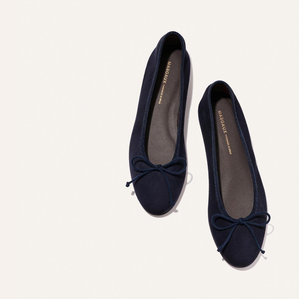 The Demi - Indigo Suede by Margaux: a pair of handmade Spanish ballet flats in dark navy blue suede, featuring small toe bows, shown from above on a light off-white background.