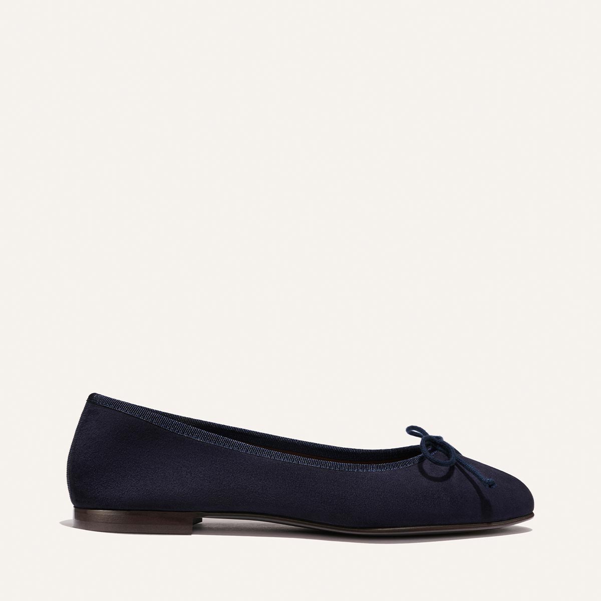 The Demi - Indigo Suede by Margaux is a handmade navy suede ballet flat with a small bow, round toe, and low brown heel, shown in side profile on a white background.