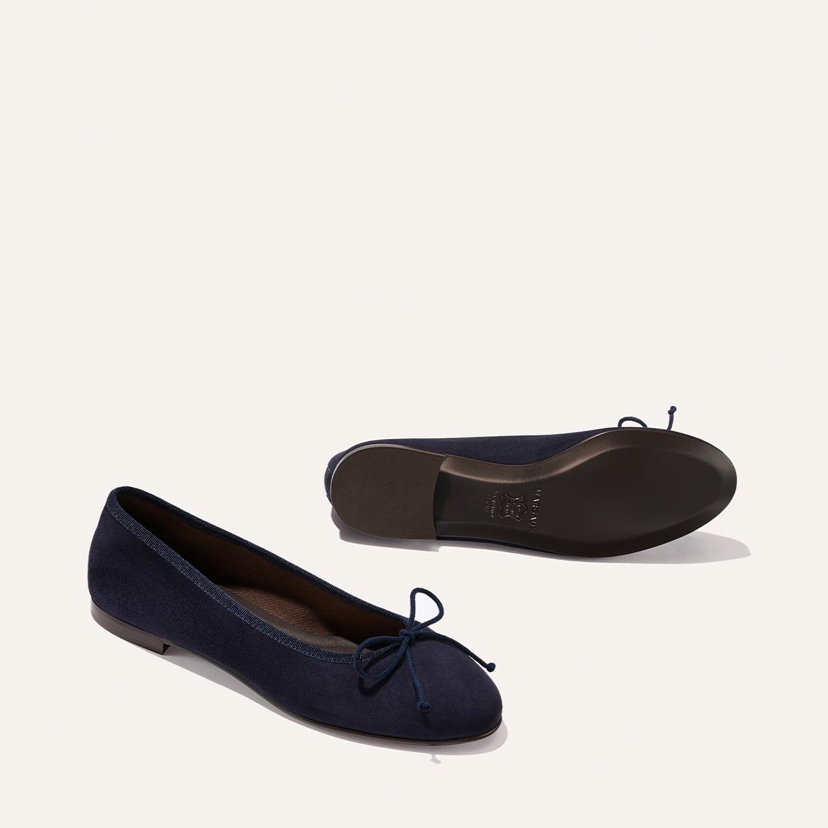 Margaux’s The Demi - Indigo Suede ballet flats, handmade in Spain, are shown on a plain background—one upright and one on its side revealing the sole. The comfortable navy blue shoes feature a small bow at the toe.