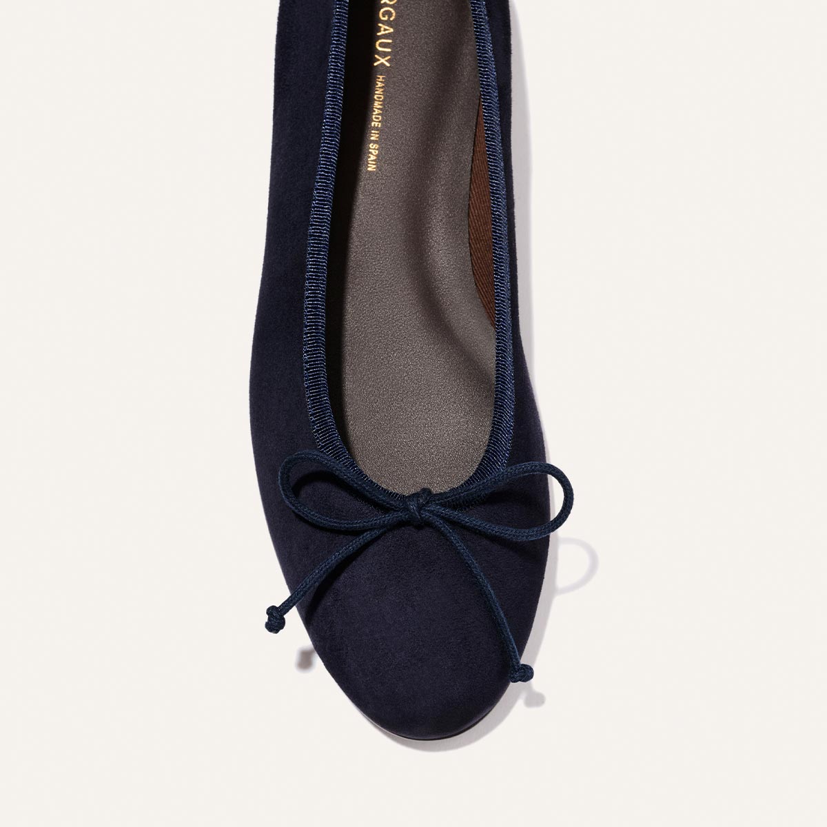 A close-up overhead view of The Demi - Indigo Suede by Margaux, a comfortable ballet flat handmade in Spain with a matching bow on the rounded toe and gold writing visible on the insole.