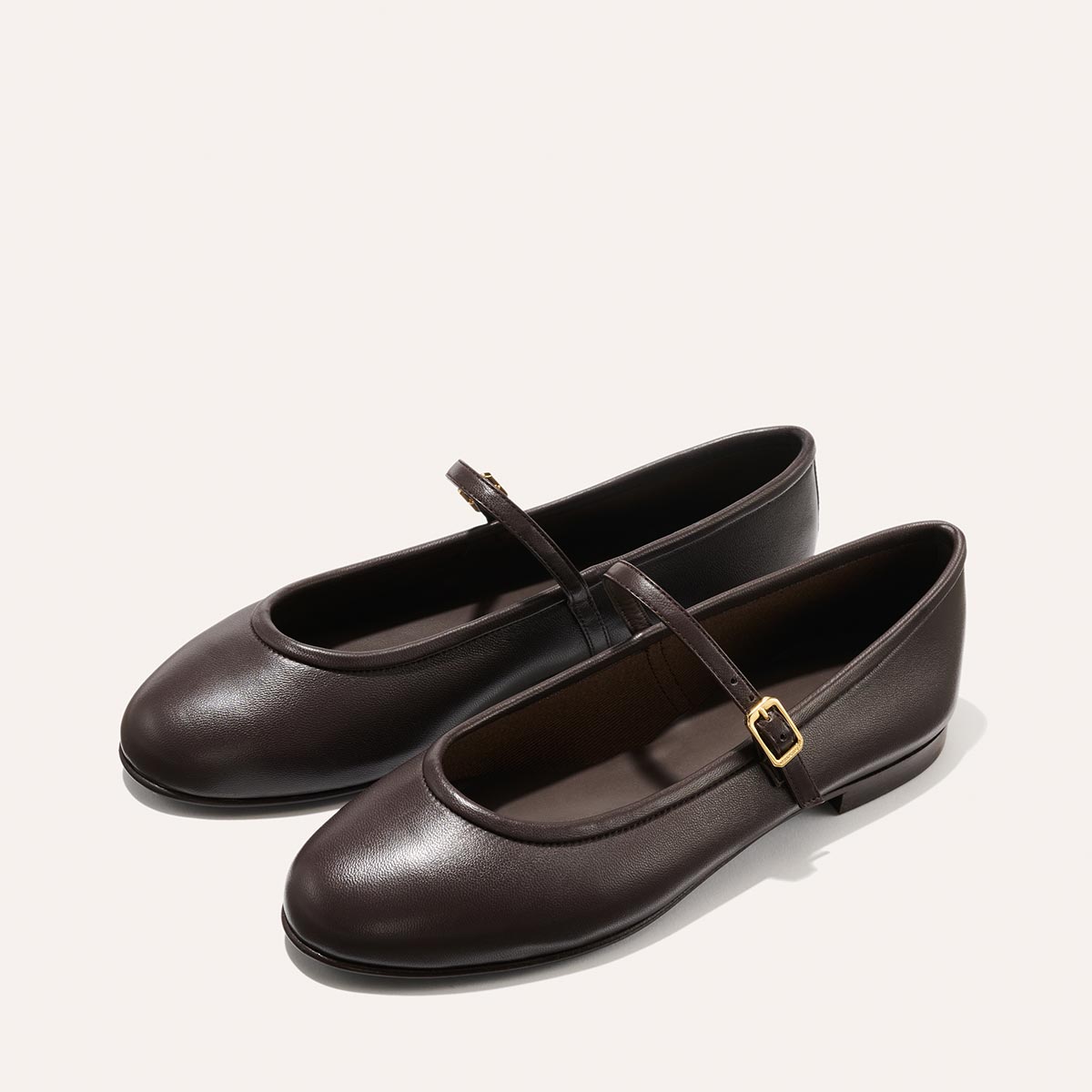 The Demi Jane - Espresso Nappa by margaux-development are elegant Mary Jane flats in rich dark brown with rounded toes and thin gold-buckled straps, showcased on a light background for a chic, feminine touch.