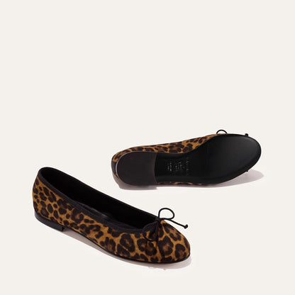 A pair of The Demi - Leopard Suede ballet flats by margaux-development, handmade in Spain, featuring leopard print suede, black trim, and small black bows. One shoe is upright and one is on its side showing the sole against a plain light background.
