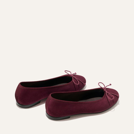 A pair of The Demi - Merlot Suede ballet flats by Margaux, handmade in Spain and adorned with petite bows on the front, are displayed on a neutral background, positioned slightly askew from each other.